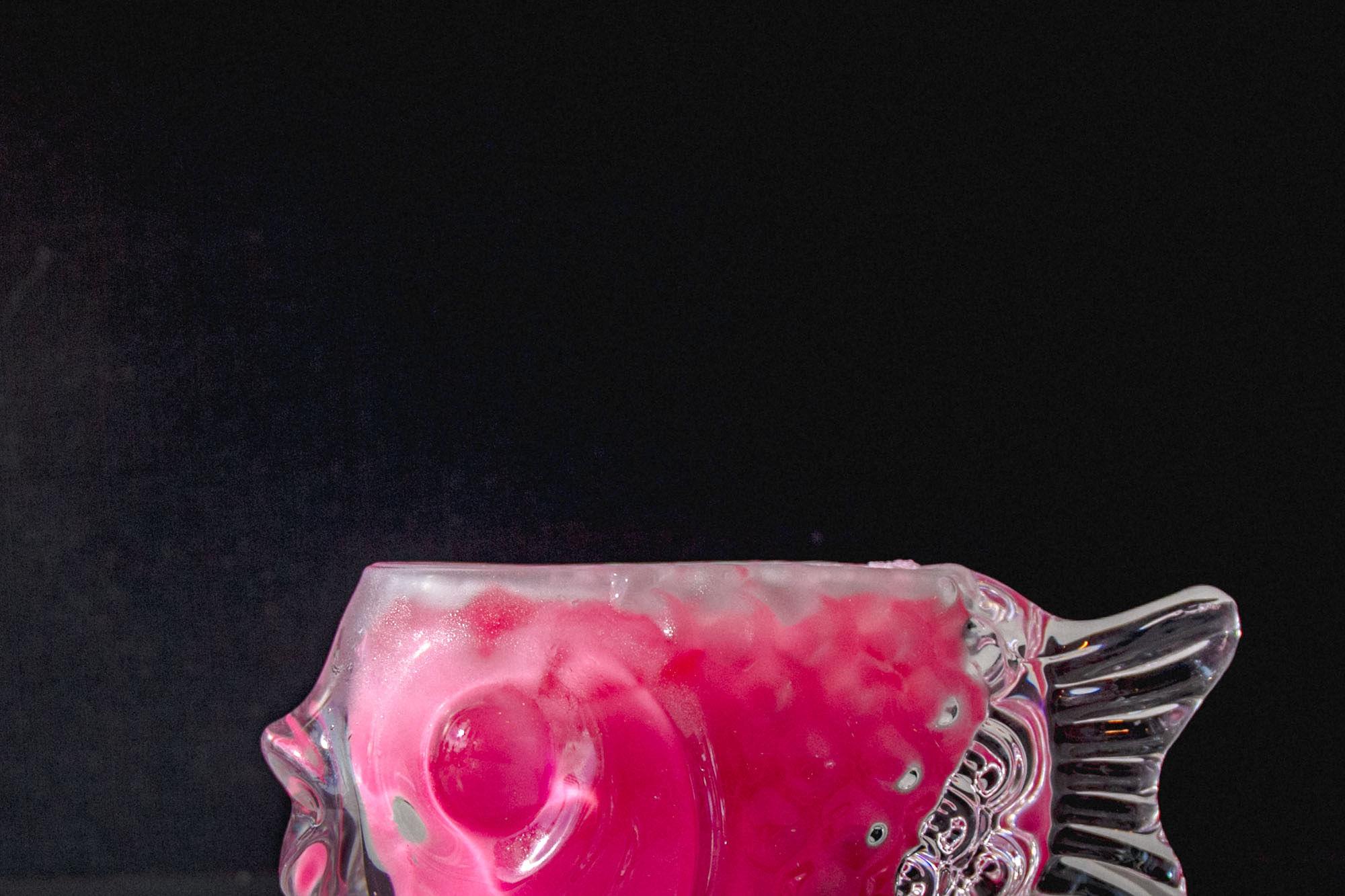 A fish-shaped glass filled with the bright pink Nom Yen drink, lit against a dark background