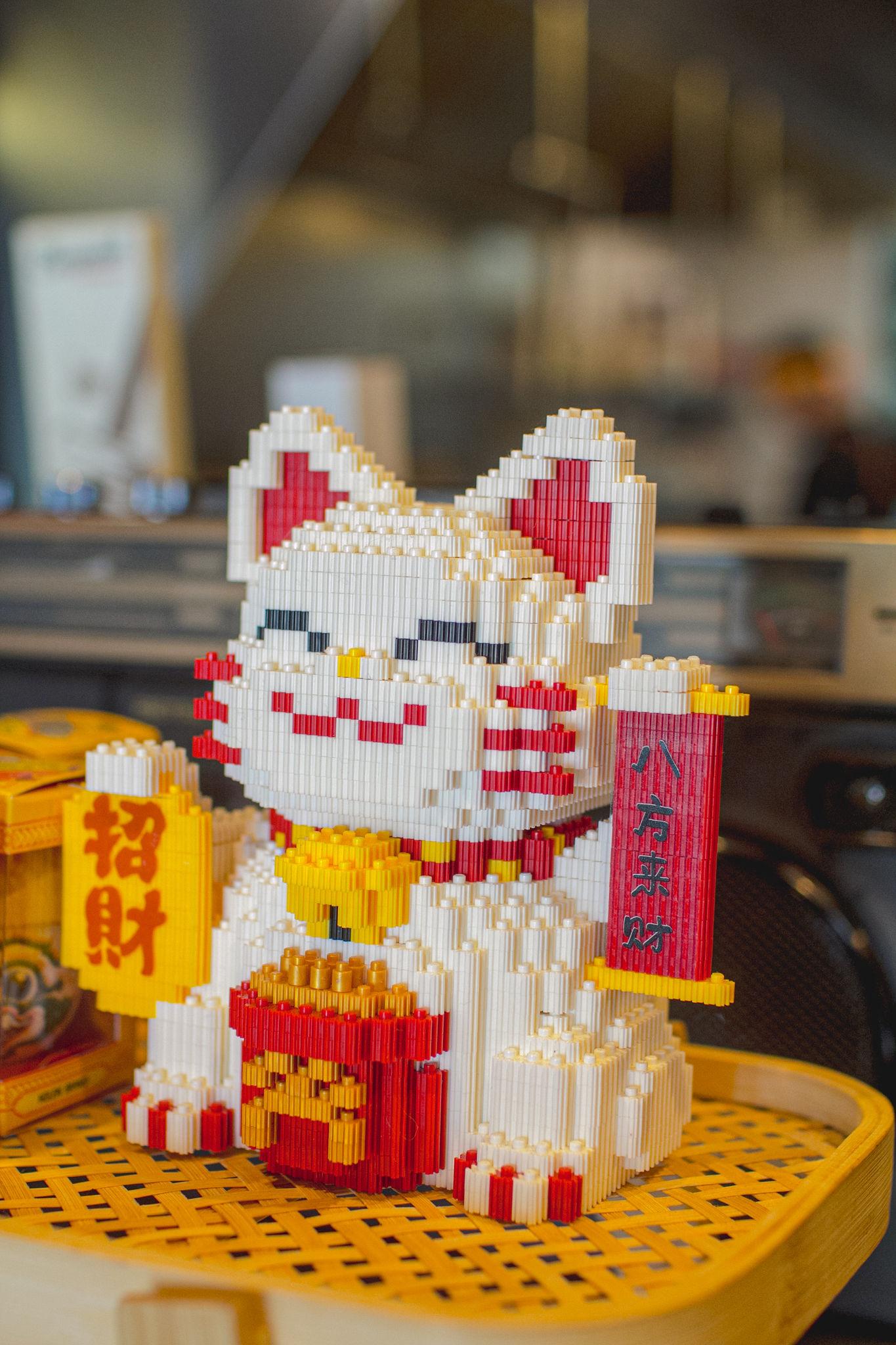 A maneki-neko lucky cat built from tiny plastic bricks, sitting on a woven bamboo tray